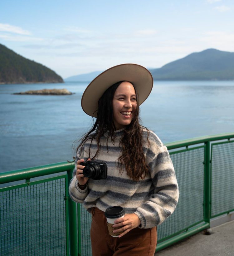 Pacific Northwest travel blogger Kara Patajo on a Washington State ferry. She is holding a Nikon z30 camera and a coffee.